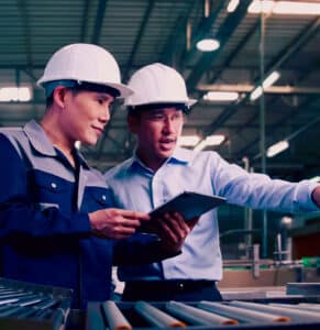 Two engineers wearing safety helmets and holding clipboards, collaborating on a Hazard and Operability (HAZOP) study in an industrial facility to identify risks and enhance safety measures.