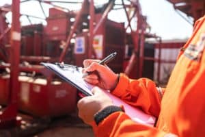 Engineer in orange protective gear conducting an incident investigation with a clipboard in an industrial facility.